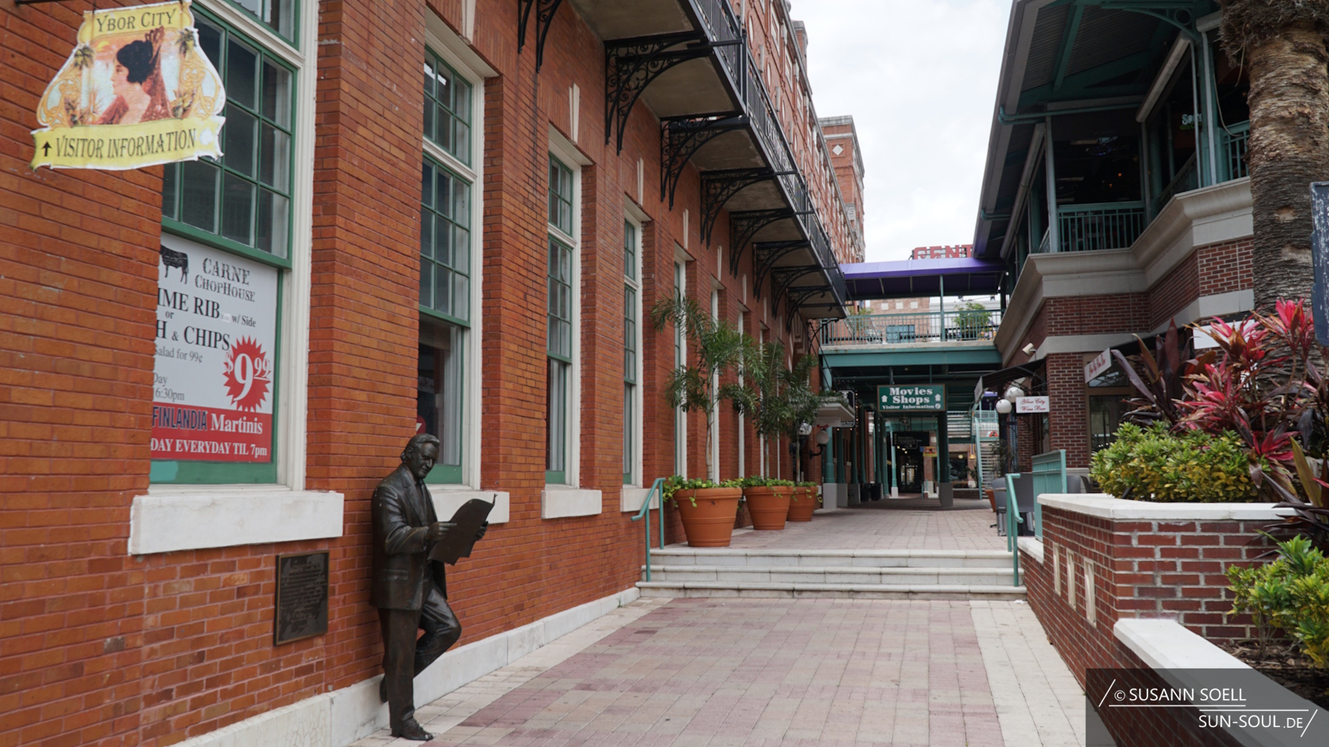 Straßenszene in Ybor City mit roten Backsteingebäuden, grünen Fensterrahmen und schmiedeeisernen Balkonen. An der linken Hauswand lehnt eine bronzene Statue eines Mannes, der eine Zeitung liest. Darüber hängt ein Schild mit der Aufschrift "Ybor City Visitor Information". Im Fenster daneben wirbt ein Plakat des "Carne Chophouse" für ein Prime‑Rib‑ und Fish‑and‑Chips‑Angebot sowie Finlandia‑Martinis. Der gepflasterte Weg führt zu einem Bereich mit dem Schild "Movies Shops".